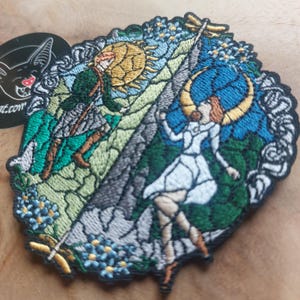 Puede incluir: Parche bordado con un dise&ntilde;o de estilo vidriera. El parche representa dos figuras, una con un sol y otra con una luna creciente, sobre un fondo de monta&ntilde;as, flores y una lib&eacute;lula. El parche tiene un borde negro.