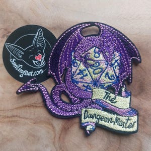 Op de afbeelding: Geborduurde patch met een paarse draak met een D20 dobbelsteen in het midden. De patch bevat de tekst "The Dungeon Master" op een banner. Er is ook een zwarte ronde sticker met een vleermuislogo aanwezig.
