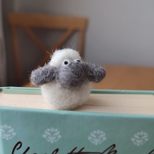 May include: A small white and gray felt sheep bookmark with black eyes. The bookmark is sitting on top of a teal book with the text "Charlotte Bronte" on the cover.