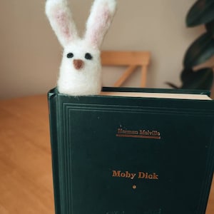 May include: A white felt bunny bookmark with black eyes and a brown nose, perched on a green book titled "Moby Dick" by Herman Melville.