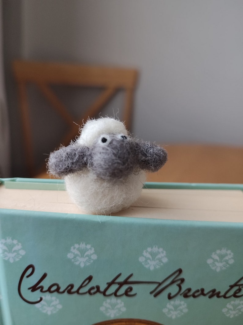 May include: A small, white and gray felt sheep bookmark. The sheep has black eyes and a white face. It is sitting on top of a teal book with the text "Charlotte Bronte" on the cover.