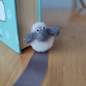May include: A white and gray felt sheep bookmark with black eyes. The bookmark is attached to a gray ribbon.