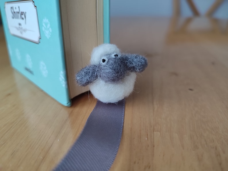 May include: A white and gray felt sheep bookmark with black eyes. The bookmark is attached to a gray ribbon.