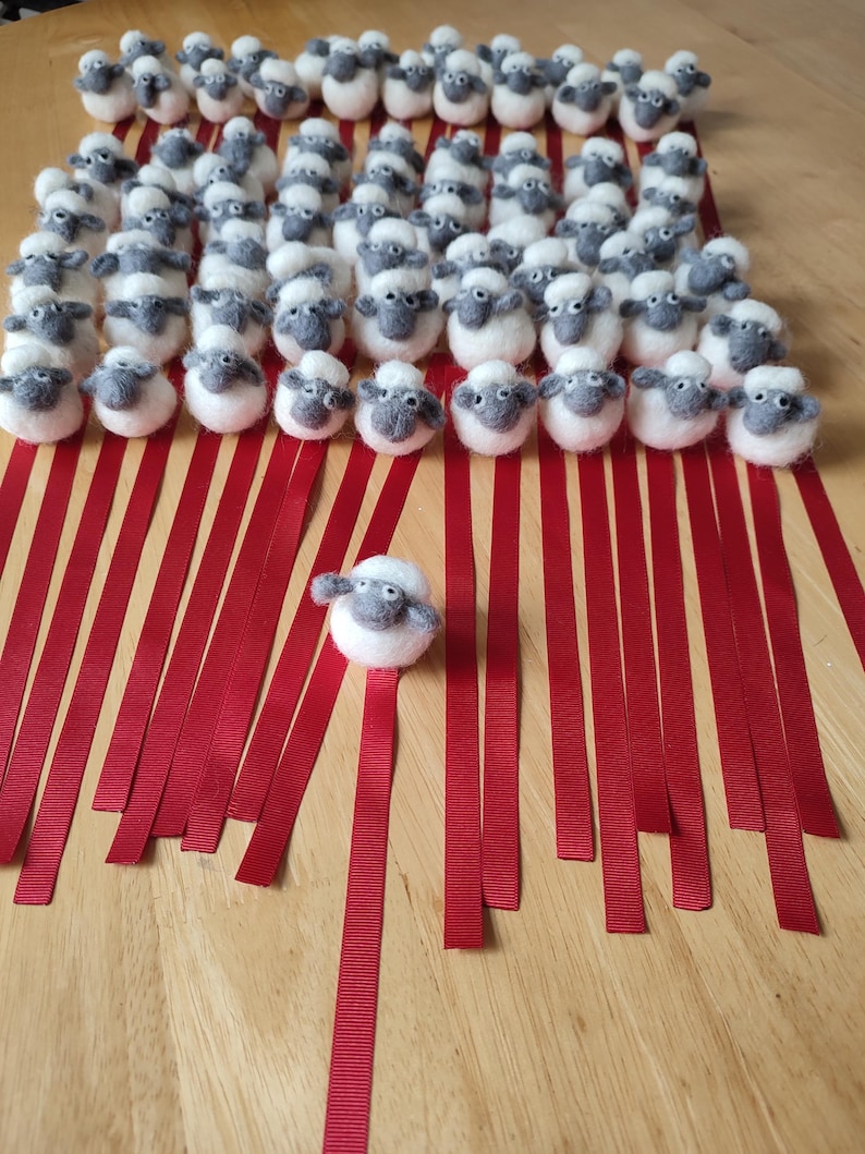 May include: A group of white felt sheep with gray faces are lined up on red ribbons. The sheep are all facing forward and have a single sheep in the center of the image.