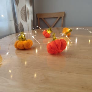 May include: A string of fairy lights with felt pumpkins in orange and yellow, arranged on a wooden table.