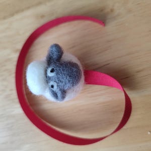 May include: A small, white and gray felt sheep with black eyes is attached to a red ribbon. The sheep is a handmade craft.