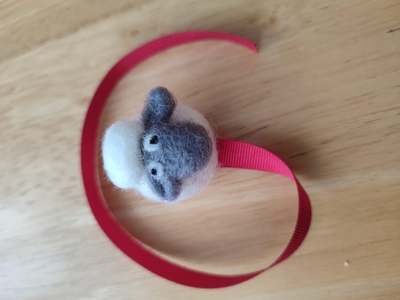 May include: A small, white and gray felt sheep with black eyes is attached to a red ribbon. The sheep is a handmade craft.
