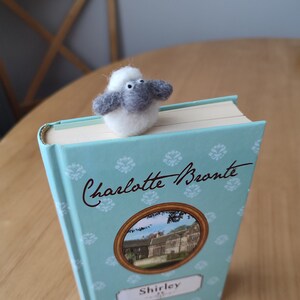 May include: A gray and white felt sheep bookmark on top of a blue book with the title "Charlotte Bronte Shirley" on the cover.