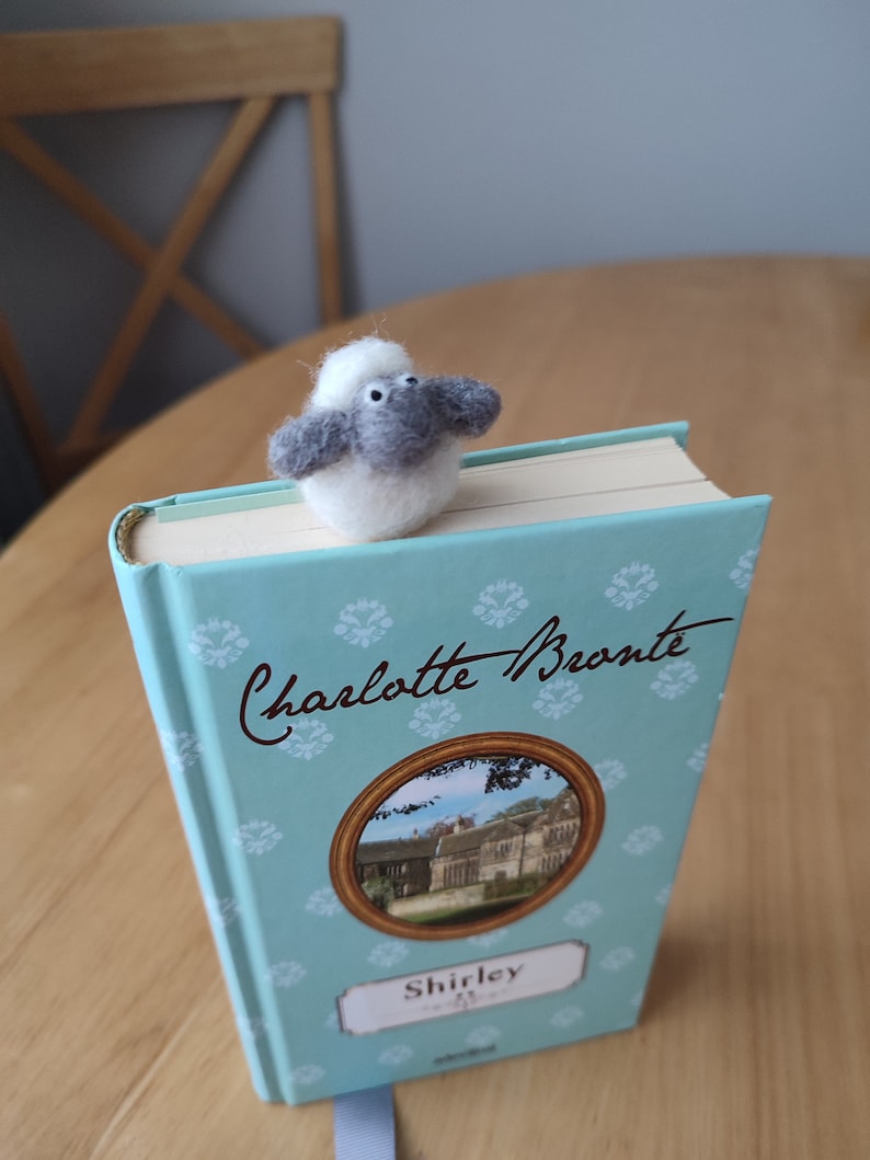 May include: A gray and white felt sheep bookmark on top of a blue book with the title "Charlotte Bronte Shirley" on the cover.