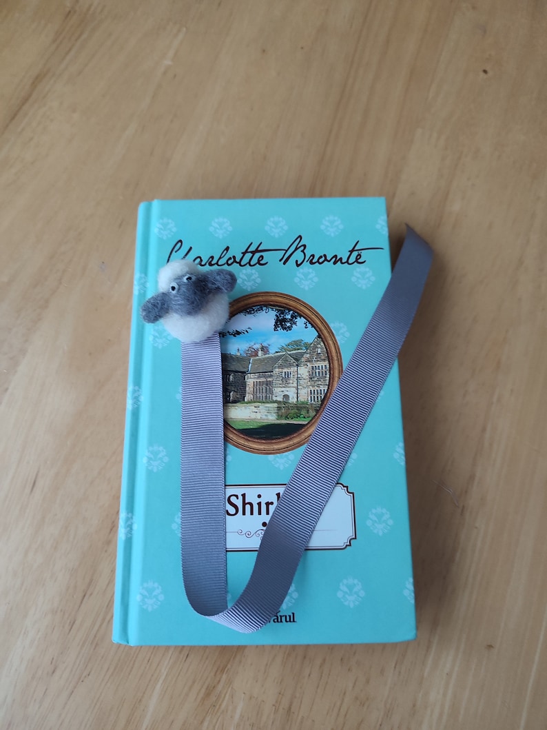May include: A teal blue book with a gray ribbon bookmark and a small gray and white sheep figurine. The book cover features the text "Charlotte Bronte" and "Shirley" with a picture of a house.