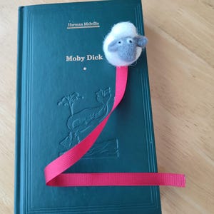 May include: A green hardcover book with the title "Moby Dick" by Herman Melville. A red ribbon bookmark with a white and gray felt sheep head is attached to the book.