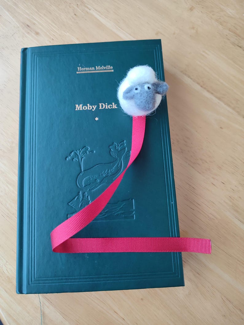 May include: A green hardcover book with the title "Moby Dick" by Herman Melville. A red ribbon bookmark with a white and gray felt sheep head is attached to the book.