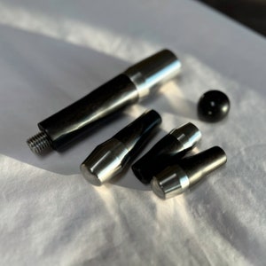 Könnte beinhalten: Eine Ansammlung von schwarzen und silberfarbenen Metallkomponenten, darunter eine Gewindestange, mehrere konische Teile und eine kleine Kugel. Die Teile sind auf einer weißen Oberfläche angeordnet, wobei der Fokus auf ihrer metallischen Oberfläche und ihrem Design liegt. Maße in cm.