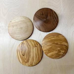 Könnte beinhalten: Vier runde Untersetzer aus Holz in verschiedenen Braun- und Beigetönen. Die Untersetzer weisen einzigartige Holzmaserungsmuster auf, wobei einer heller und die anderen dunkler sind. Die Untersetzer sind auf einer hellen Holzoberfläche angeordnet.