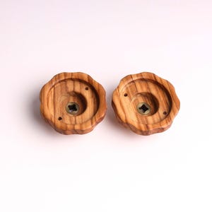 Puede incluir: Dos pomos de madera con forma circular y un agujero en el centro. Los pomos tienen un patrón de veta de madera natural y están hechos de madera marrón claro.