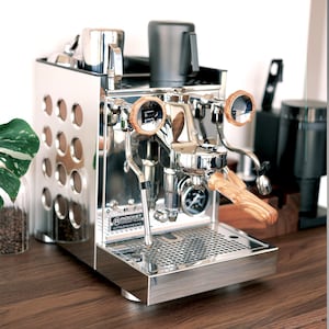 May include: A silver Rocket Appartamento espresso machine with wood accents. The machine has a stainless steel body with a perforated drip tray. The machine has two portafilters with wooden handles.