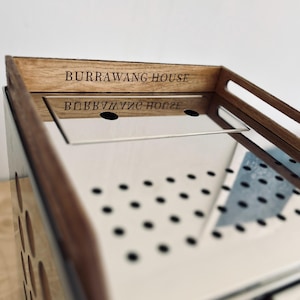 May include: A stainless steel and wood serving tray with the text "BURRAWANG HOUSE" engraved on the wooden frame. The tray has a mirrored surface and a perforated design on the sides. The tray has a rectangular shape.