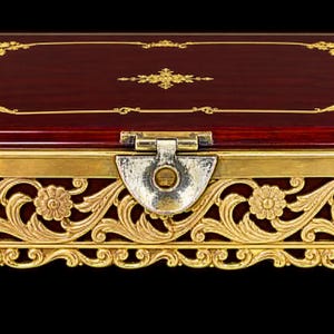 May include: A rectangular wooden box with a deep mahogany finish and a polished surface. The lid features a gold-colored floral border and central motif. The base is encased in an ornate gold-toned metal filigree with floral accents.