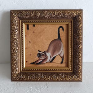 Pode incluir: Pintura emoldurada de um gato siamês a alongar-se. O gato tem um corpo bege, pontas castanhas escuras e olhos azuis. A obra de arte está numa moldura dourada ornamentada, com um fundo bege e pegadas.