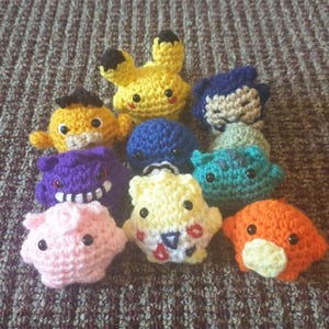 May include: A collection of handmade crochet figures in various colors, including yellow, blue, purple, pink, and orange. Each figure has a unique design with black eyes and detailed features. The figures are arranged on a textured surface.