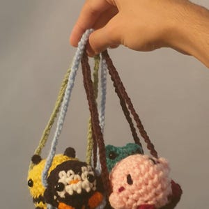 May include: Three crocheted plushie keychains hanging from their straps. The plushies are a bee, a frog, and a pink character. The bee is yellow and black with a white flower on its head. The frog is green and the pink character has a red blush on its cheeks.