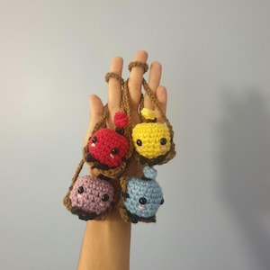 May include: Four crocheted plushie keychains hanging from a hand. The keychains are shaped like small bags with faces. The bags are red, yellow, purple, and blue.