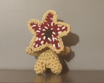 May include: A crocheted plush toy shaped like a monster with a red and white star-shaped head. The toy is beige and has a brown mouth.