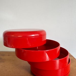 May include: A bright red, multi-tiered circular storage container with a glossy finish. The object consists of four stacked, round, shallow trays that are offset from one another, creating a stepped, spiral-like appearance with a solid lid.