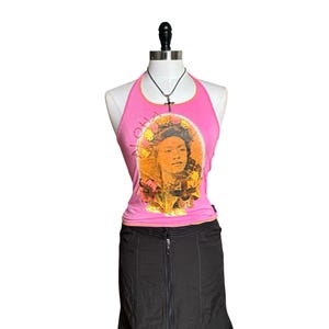 May include: A pink halter top featuring a portrait of a woman encircled by flowers and the word "Aloha". The top is worn with a black skirt. A black necklace with a cross pendant is also visible.