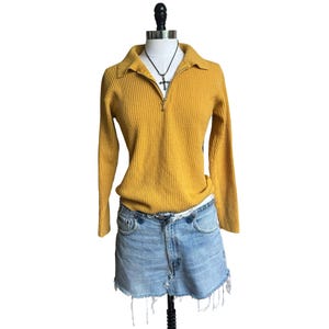 May include: A mustard yellow long-sleeve ribbed knit sweater featuring a quarter-zip collar and a silver-toned cross pendant necklace, paired with a light-wash blue denim mini skirt that has a frayed, raw-edge hemline and a classic five-pocket design.