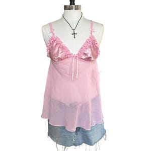 May include: A light pink camisole featuring satin cups with ruffled trim, thin shoulder straps with small bows, and a sheer mesh body. A thin ribbon tie is centered at the neckline. The garment is displayed on a mannequin wearing a cross necklace.