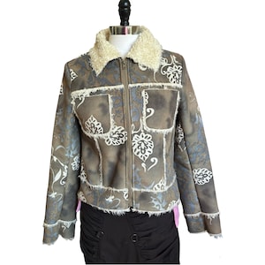 Puede incluir: Una chaqueta marrón con cuello y ribetes de piel sintética color crema. La chaqueta presenta una base marrón descolorida y moteada con un estampado floral blanco, cremallera frontal completa, dos bolsillos en el pecho y mangas largas deshilachadas.