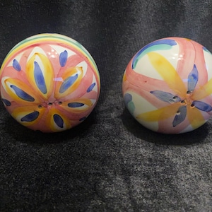 May include: Two spherical ceramic salt and pepper shakers, hand-painted with a floral design. The design features pink, yellow, and blue brushstrokes on a white base. Each shaker has multiple holes for dispensing.