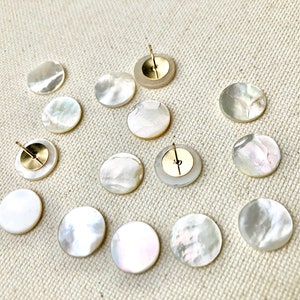 May include: Twelve white mother of pearl round buttons with gold metal backs. The buttons are arranged in a circle on a white fabric background.