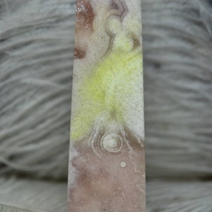 May include: A rectangular crystal obelisk with a pointed top. The stone features a blend of colors, including pale pink, yellow, and white, with circular patterns. The letter "B" is in a black square in the upper left corner.
