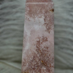 May include: A pink and white crystal obelisk with a pointed top. The stone has a mottled appearance with areas of white and darker pink. The obelisk is a rectangular shape with a flat base.