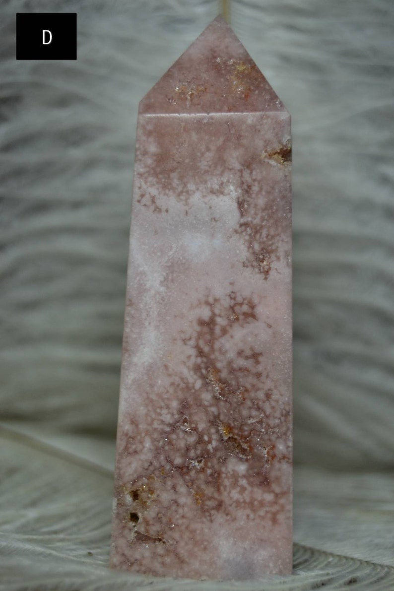 May include: A pink and white crystal obelisk with a pointed top. The stone has a mottled appearance with areas of white and darker pink. The obelisk is a rectangular shape with a flat base.