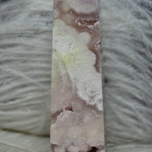 May include: A tall, rectangular crystal point with a light purple hue and white, cloud-like inclusions. The crystal has a polished surface and a rough, geode-like cavity near the base. The background is a soft, white, feathery texture.