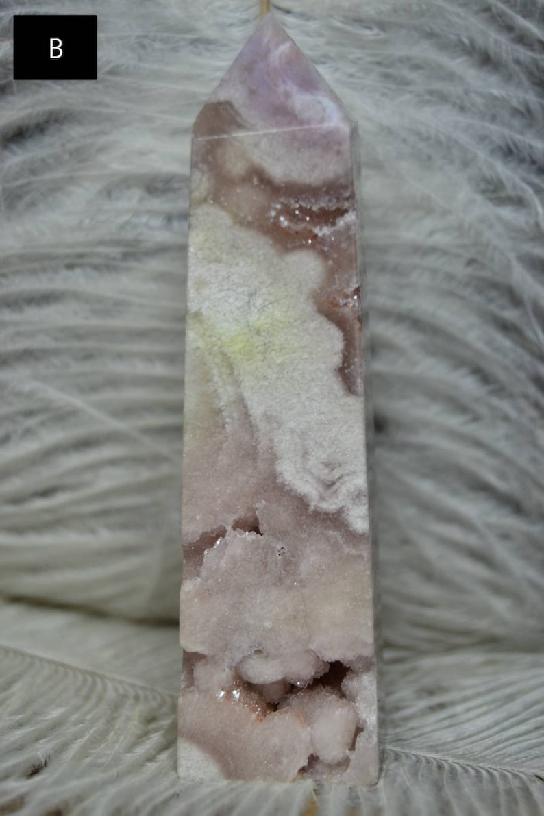 May include: A tall, rectangular crystal point with a light purple hue and white, cloud-like inclusions. The crystal has a polished surface and a rough, geode-like cavity near the base. The background is a soft, white, feathery texture.