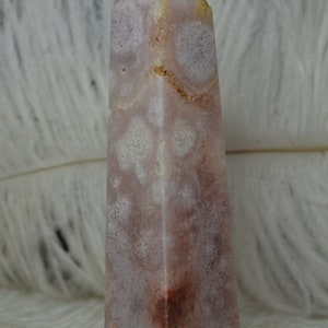 May include: A tall, rectangular crystal obelisk with a pointed top. The stone features a mix of pale pink, white, and brown hues, with a speckled pattern. The background is a soft, white, feathery texture.