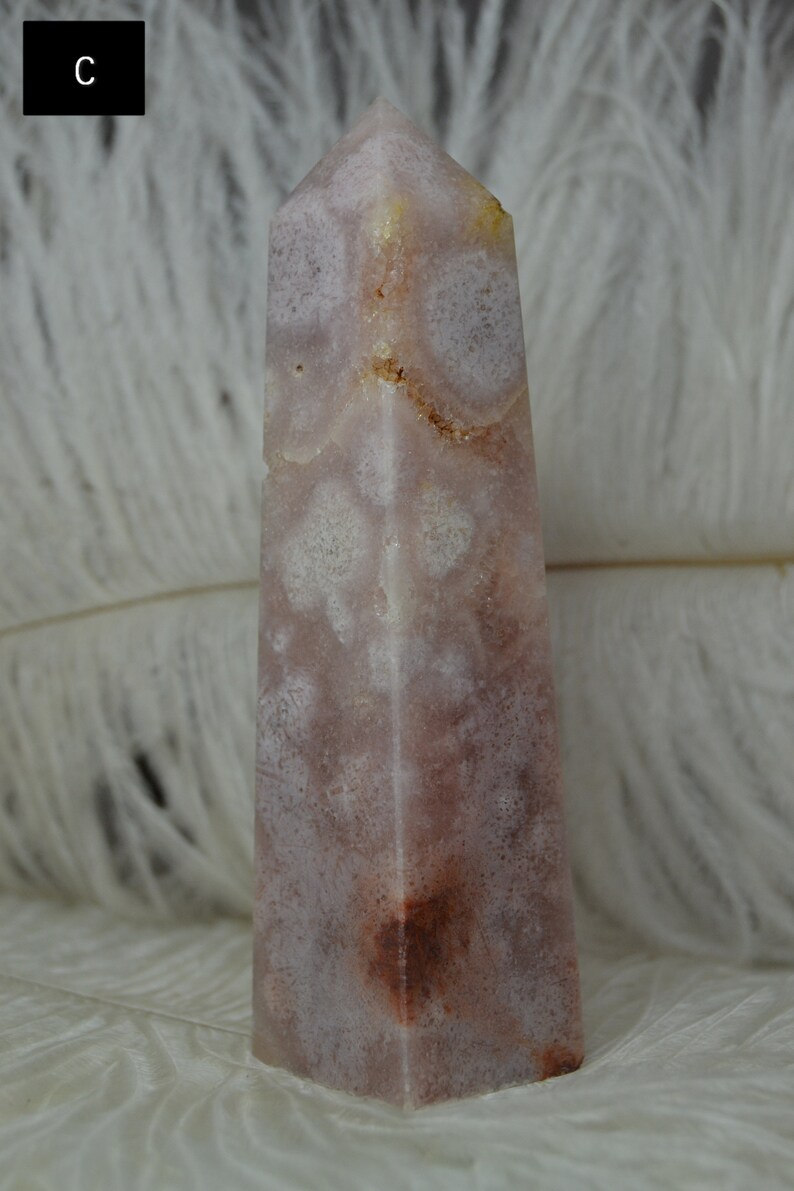 May include: A tall, rectangular crystal obelisk with a pointed top. The stone features a mix of pale pink, white, and brown hues, with a speckled pattern. The background is a soft, white, feathery texture.