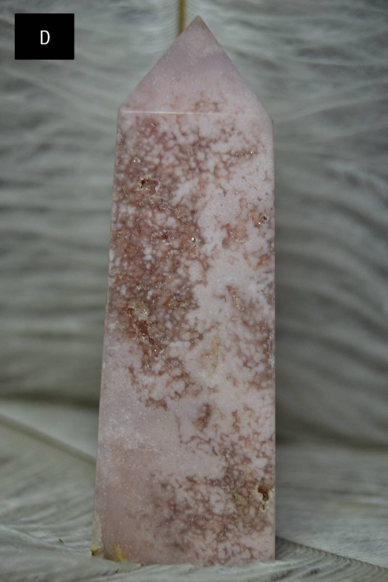 May include: A pink and brown crystal tower with a pointed top. The crystal has a unique pattern of brown and white veining throughout. The tower is a decorative item, likely used for display or spiritual purposes.