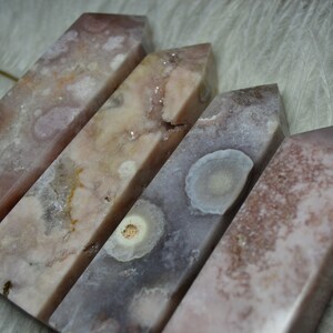 May include: Four polished, rectangular crystals with angled tops. The crystals display a mix of pink, white, and gray hues with unique patterns. The crystals are arranged diagonally, showcasing their varying textures and colors.