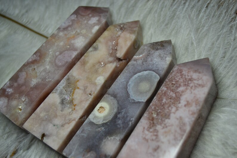 May include: Four polished, rectangular crystals with angled tops. The crystals display a mix of pink, white, and gray hues with unique patterns. The crystals are arranged diagonally, showcasing their varying textures and colors.