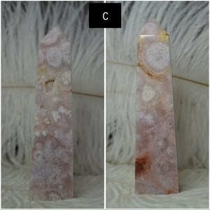 May include: A pink and white crystal obelisk with a pointed top. The stone has a natural pattern of white and pink swirls and spots. The obelisk is standing upright against a white, feathery background. The letter "C" is in a black box in the upper left corner.
