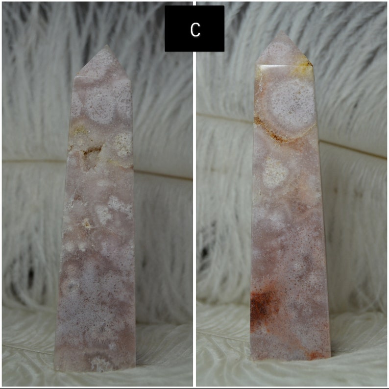 May include: A pink and white crystal obelisk with a pointed top. The stone has a natural pattern of white and pink swirls and spots. The obelisk is standing upright against a white, feathery background. The letter "C" is in a black box in the upper left corner.