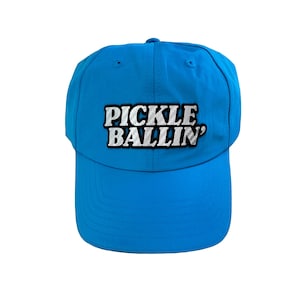 May include: A vibrant blue baseball cap featuring the text "PICKLE BALLIN'" in white and silver. The cap has a curved peak and a classic six-panel design, suitable for outdoor activities or casual wear.
