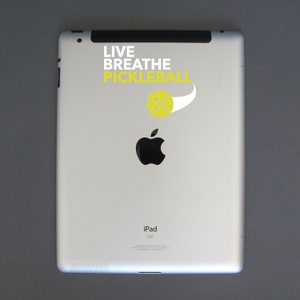 K&ouml;nnte beinhalten: Wei&szlig;es iPad mit einem schwarzen Apple-Logo und dem Text "LIVE BREATHE PICKLEBALL" mit einer gelben Pickleball-Grafik. Das iPad befindet sich auf einer grauen Oberfl&auml;che.