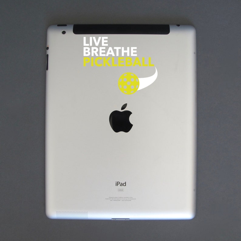 K&ouml;nnte beinhalten: Wei&szlig;es iPad mit einem schwarzen Apple-Logo und dem Text "LIVE BREATHE PICKLEBALL" mit einer gelben Pickleball-Grafik. Das iPad befindet sich auf einer grauen Oberfl&auml;che.