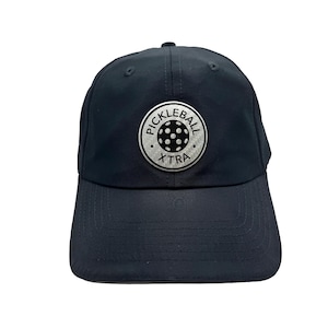May include: A navy blue baseball cap with a curved peak. The cap has a circular patch with the words "PICKLEBALL XTRA" around a pickleball graphic. The cap is made of a smooth, durable material.